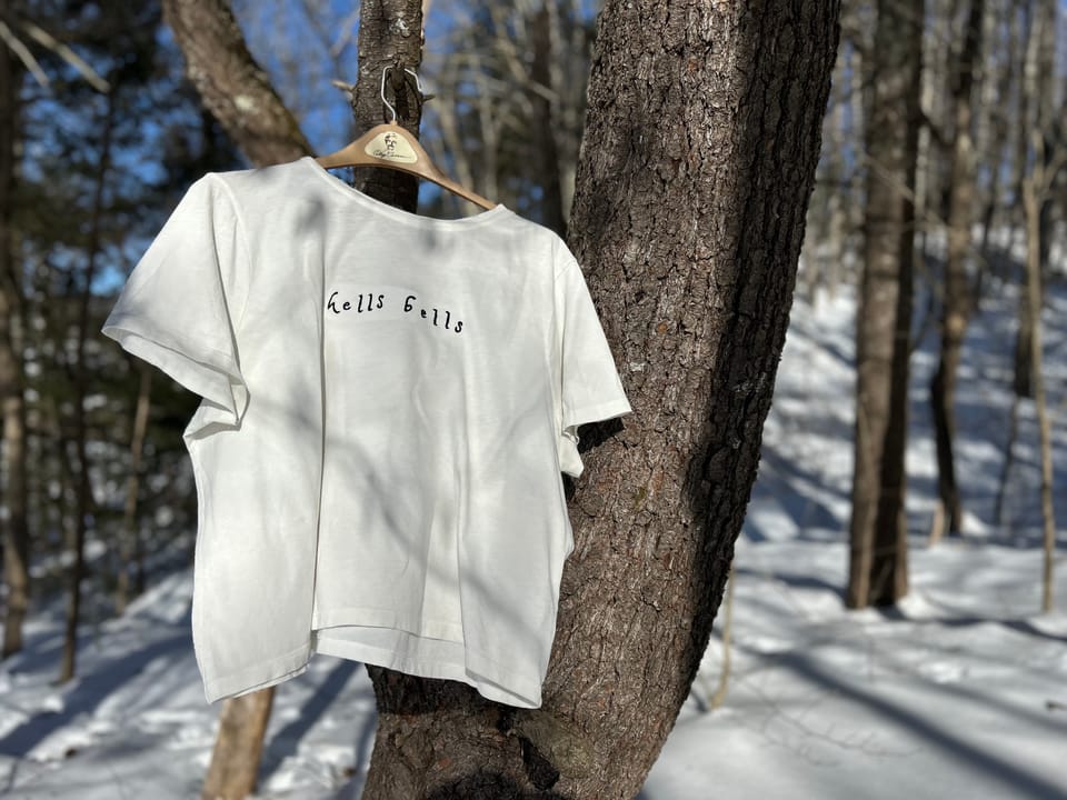 A white t-shirt with black embroidery spelling "hells bells" hanging on a tree in a forest with snow on the ground.